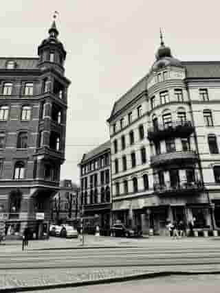 Schwarz-weiß Bild von historischen Gebäuden mit Turm und Kuppel an einer belebten Straßenecke, Personen überqueren die Straße und Autos fahren vorbei.