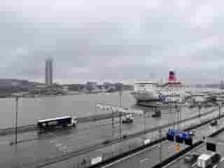 Blick auf eine Hafenszene an einem regnerischen Tag mit einem nebligen Hintergrund. Ein Schiff und mehrere Lastwagen sind sichtbar, und im Hintergrund ragt ein hohes Gebäude aus dem Nebel heraus.