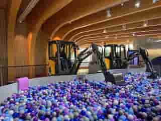 Zwei Bagger in einem Indoor-Spielbereich, gefüllt mit blauen und violetten Bällen, in einer Halle mit gewölbten Holzdecken.