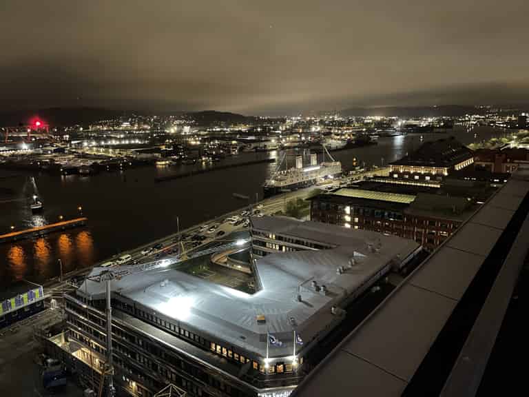 Nachtansicht eines beleuchteten Hafens mit einer leuchtenden Gebäudefassade im Vordergrund und der beleuchteten Stadt im Hintergrund, reflektiert auf dem Wasser.