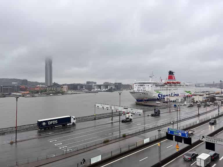 Blick auf eine Hafenszene an einem regnerischen Tag mit einem nebligen Hintergrund. Ein Schiff und mehrere Lastwagen sind sichtbar, und im Hintergrund ragt ein hohes Gebäude aus dem Nebel heraus.