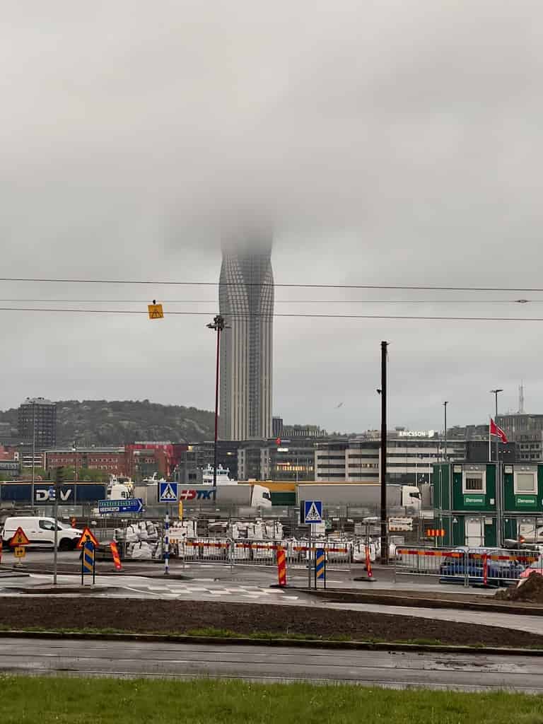 Ein hohes, schlankes Gebäude, teilweise von Nebel umhüllt, steht hinter einer Baustelle und einer verkehrsreichen Straße in einer städtischen Umgebung.