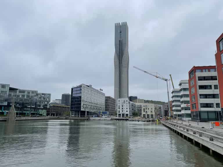 Blick auf das Ufer eines Flusses mit modernen Gebäuden und einem hohen, schlanken Turm. Das Bild zeigt eine städtische Umgebung mit einer reflektierenden Wasseroberfläche.