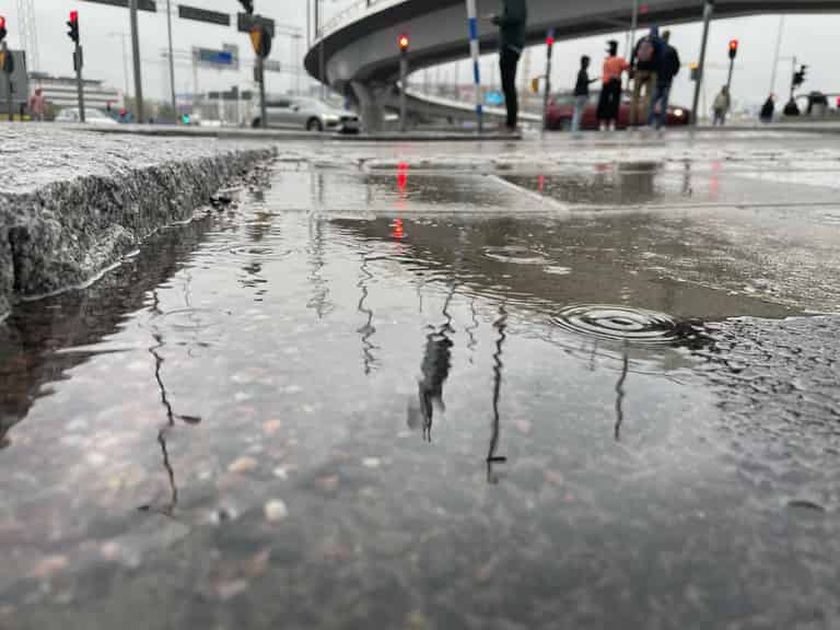 Eine städtische Szene an einem regnerischen Tag zeigt Pfützen auf dem Boden, die die Umgebung und Passanten spiegeln.