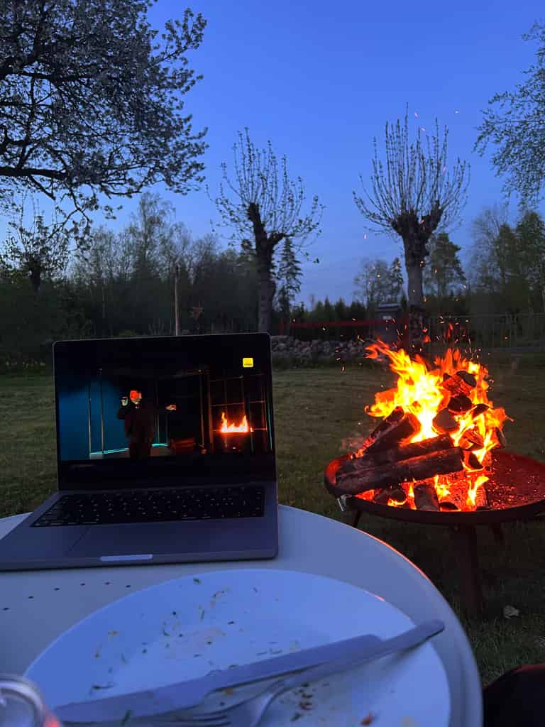 Ein Laptop, der im Freien auf einem Tisch steht, zeigt eine TV-Sendung, während ein Lagerfeuer im Hintergrund brennt.