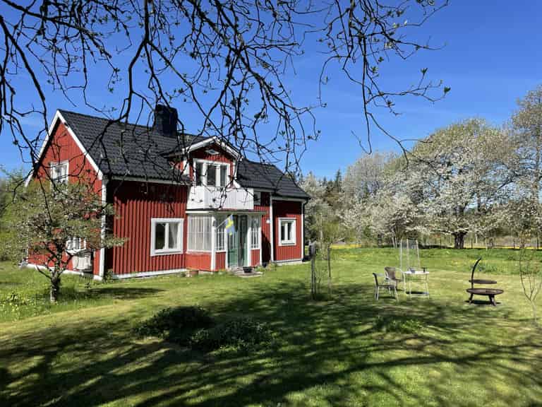 Ein traditionelles rotes schwedisches Haus mit weißer Veranda und blühenden Bäumen im Garten.