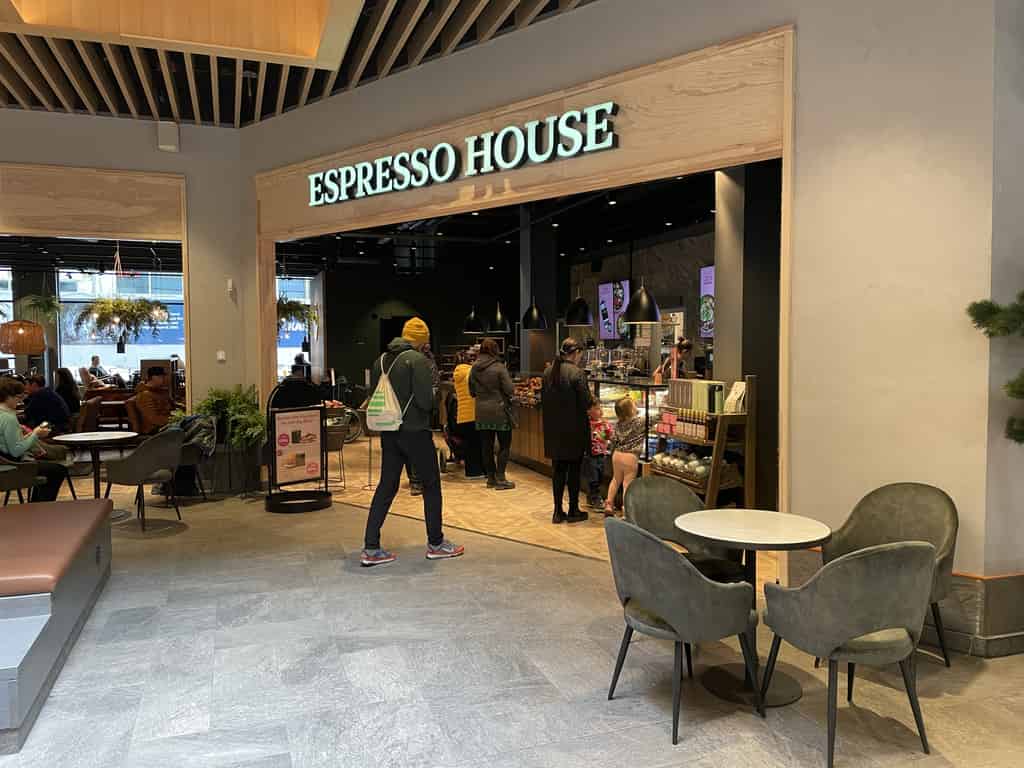 Innenansicht eines modernen Cafés mit Kunden am Tresen und anderen sitzenden Personen, umgeben von zeitgemäßer Einrichtung.
