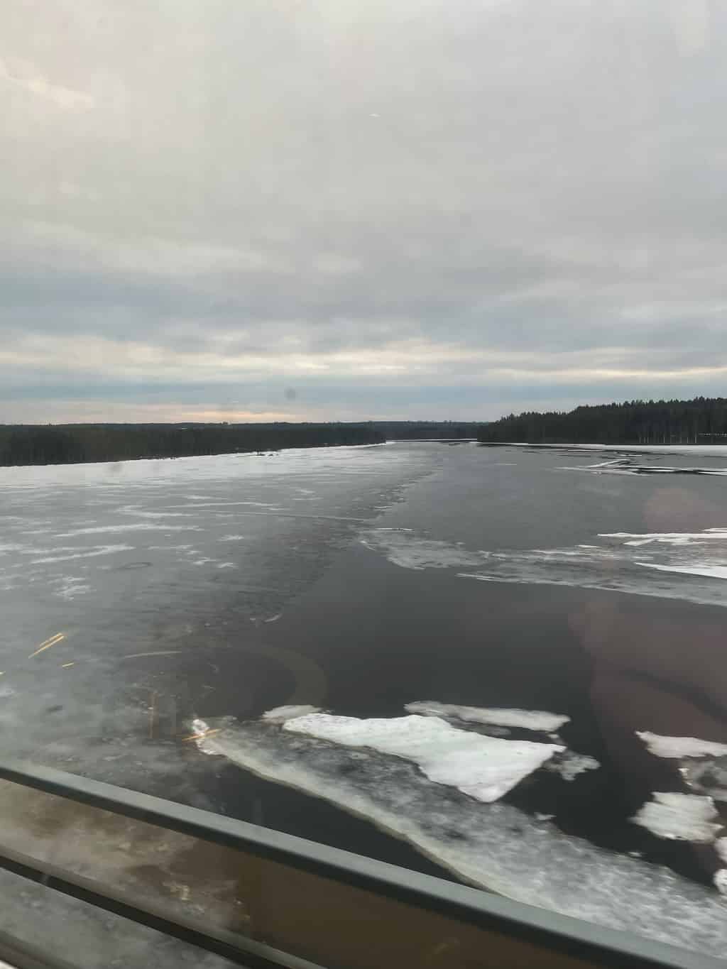 Blick aus einem Zugfenster auf einen zugefrorenen See, umgeben von bewaldeten Gebieten unter bewölktem Himmel.