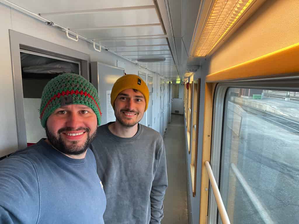 Zwei lächelnde Männer mit Strickmützen im Inneren eines Zuges, die für ein Selfie in einem gut beleuchteten Korridor posieren.