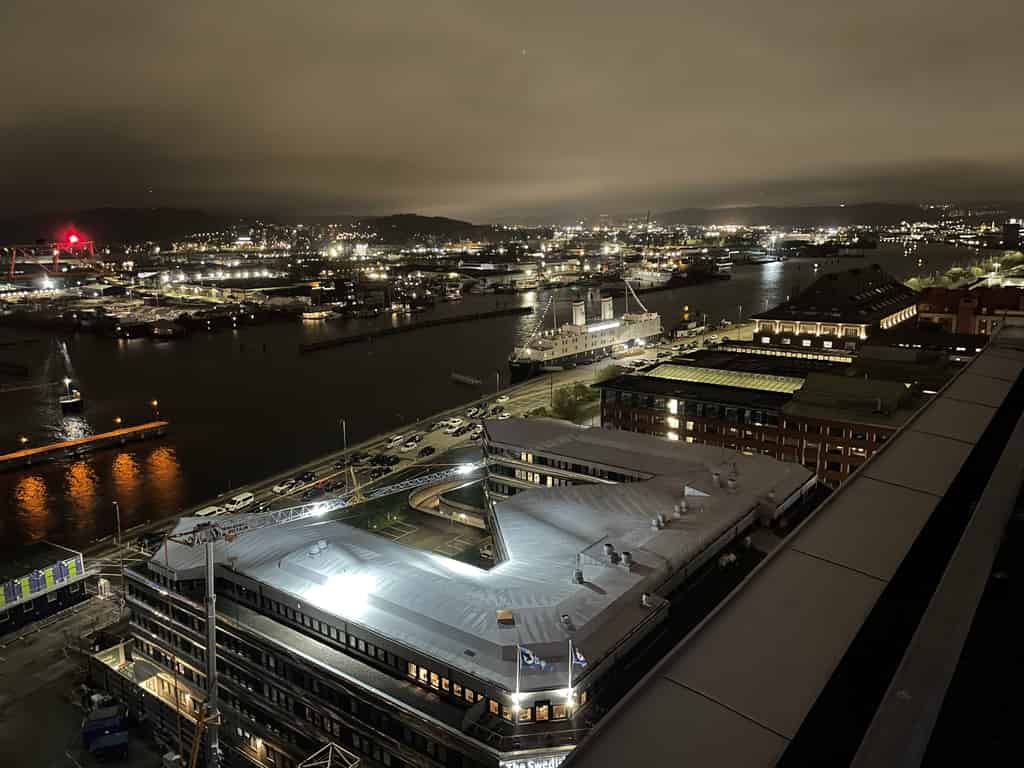 Nachtansicht eines beleuchteten Hafens mit einer leuchtenden Gebäudefassade im Vordergrund und der beleuchteten Stadt im Hintergrund, reflektiert auf dem Wasser.