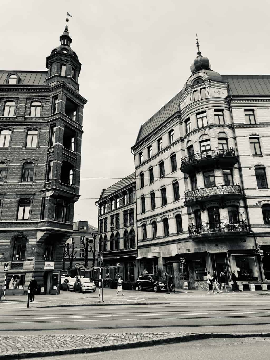 Schwarz-weiß Bild von historischen Gebäuden mit Turm und Kuppel an einer belebten Straßenecke, Personen überqueren die Straße und Autos fahren vorbei.