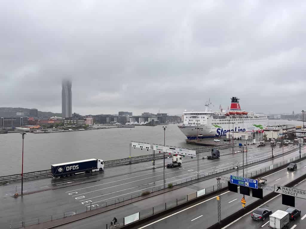 Blick auf eine Hafenszene an einem regnerischen Tag mit einem nebligen Hintergrund. Ein Schiff und mehrere Lastwagen sind sichtbar, und im Hintergrund ragt ein hohes Gebäude aus dem Nebel heraus.