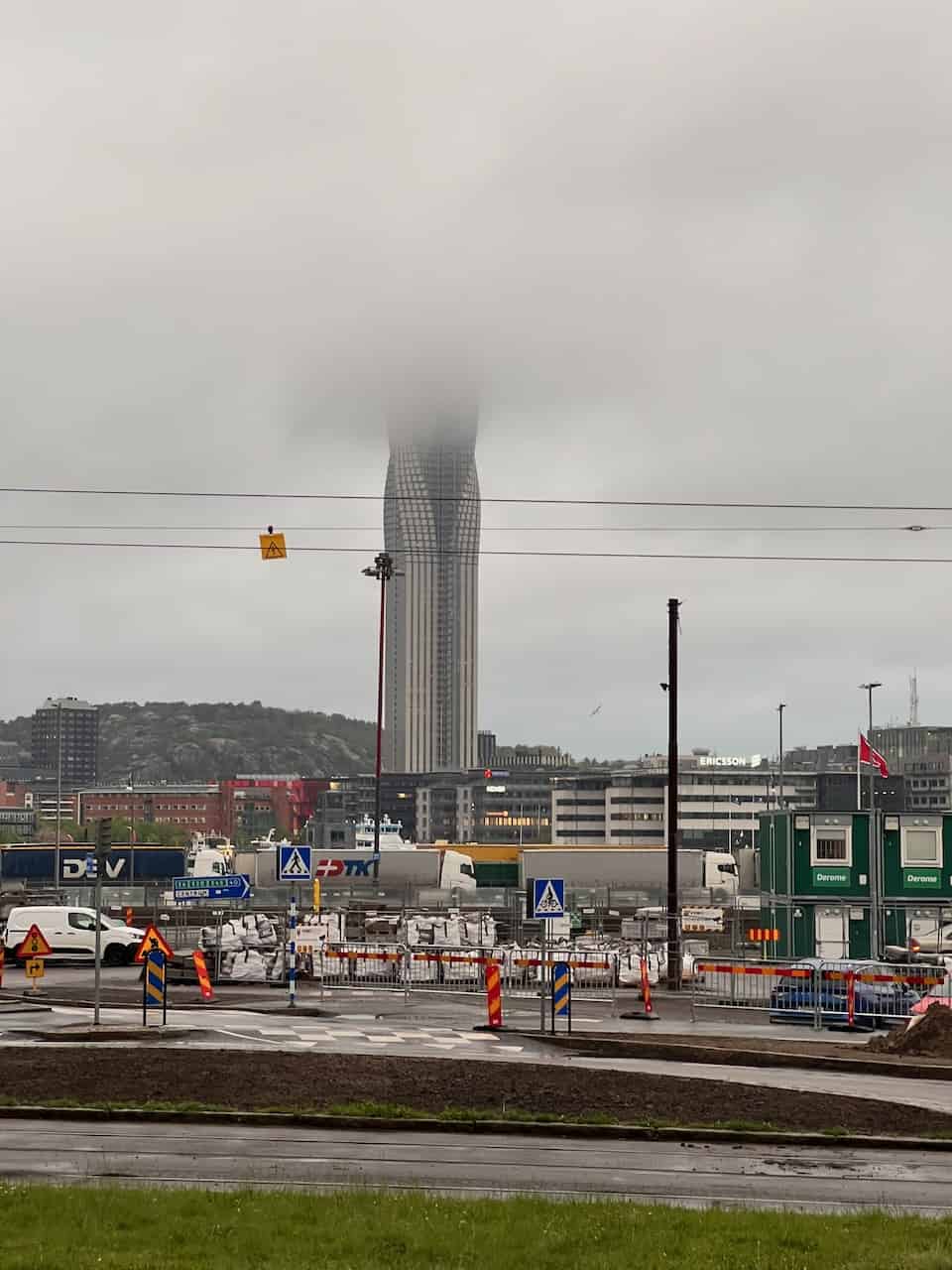 Ein hohes, schlankes Gebäude, teilweise von Nebel umhüllt, steht hinter einer Baustelle und einer verkehrsreichen Straße in einer städtischen Umgebung.