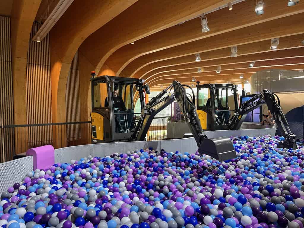 Zwei Bagger in einem Indoor-Spielbereich, gefüllt mit blauen und violetten Bällen, in einer Halle mit gewölbten Holzdecken.