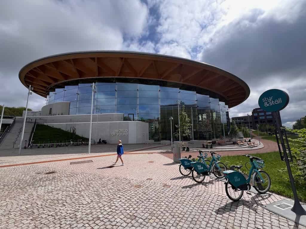 Das Gebäude 'World of Volvo' mit einer großen, kreisförmigen Holzstruktur und großen Glasfronten unter einem bewölkten Himmel. Im Vordergrund sind mehrere Mietscooter geparkt.