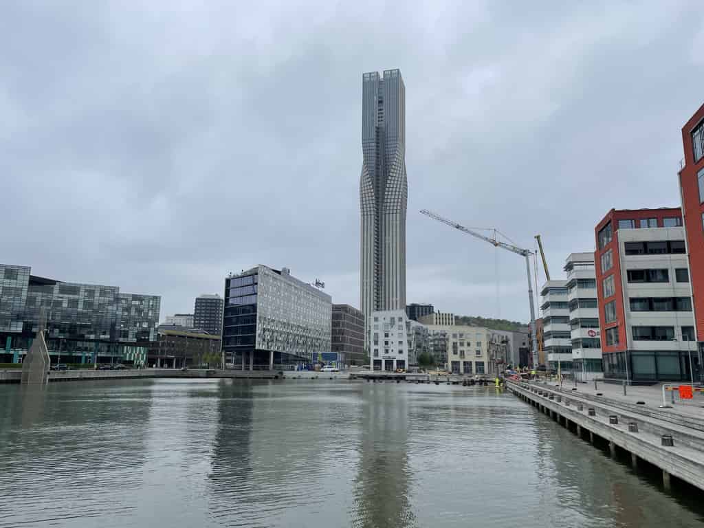 Blick auf das Ufer eines Flusses mit modernen Gebäuden und einem hohen, schlanken Turm. Das Bild zeigt eine städtische Umgebung mit einer reflektierenden Wasseroberfläche.