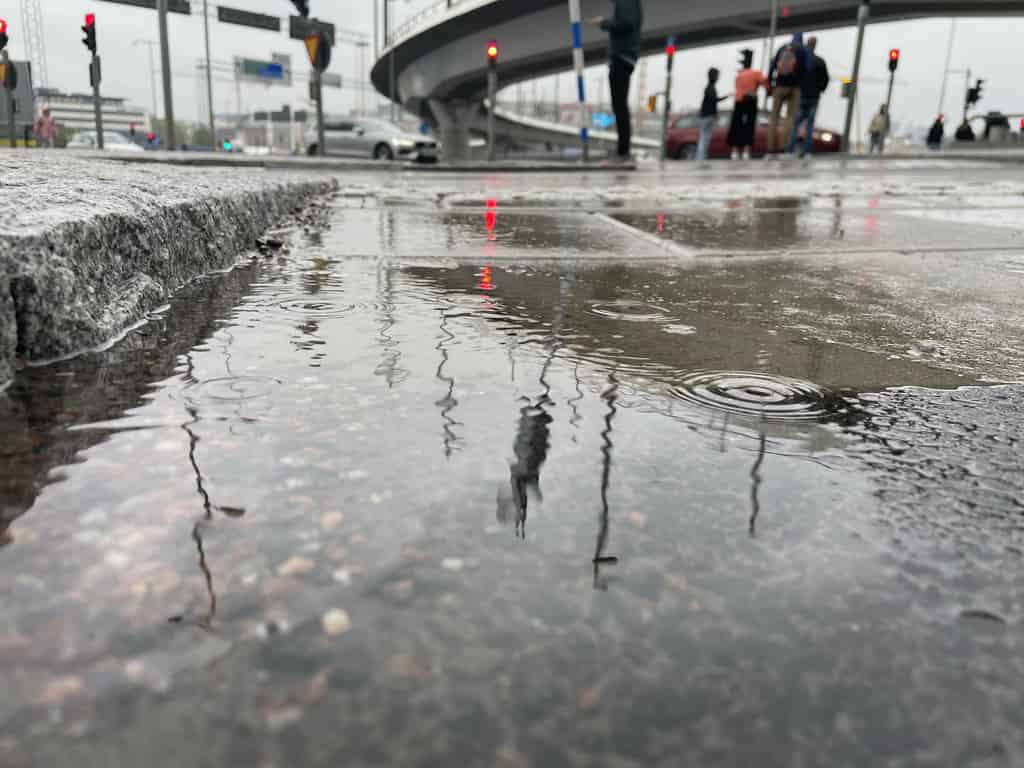 Eine städtische Szene an einem regnerischen Tag zeigt Pfützen auf dem Boden, die die Umgebung und Passanten spiegeln.