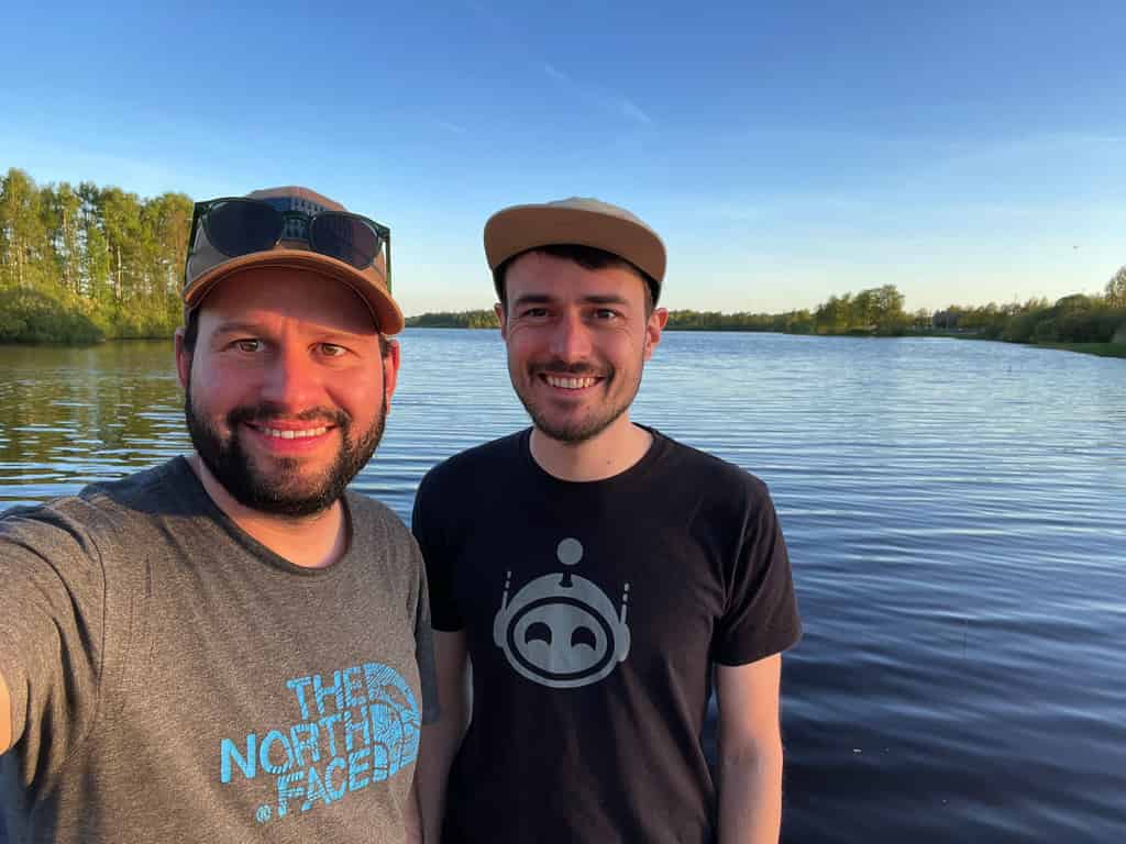Zwei Männer, die vor einem See bei Sonnenuntergang lächeln.