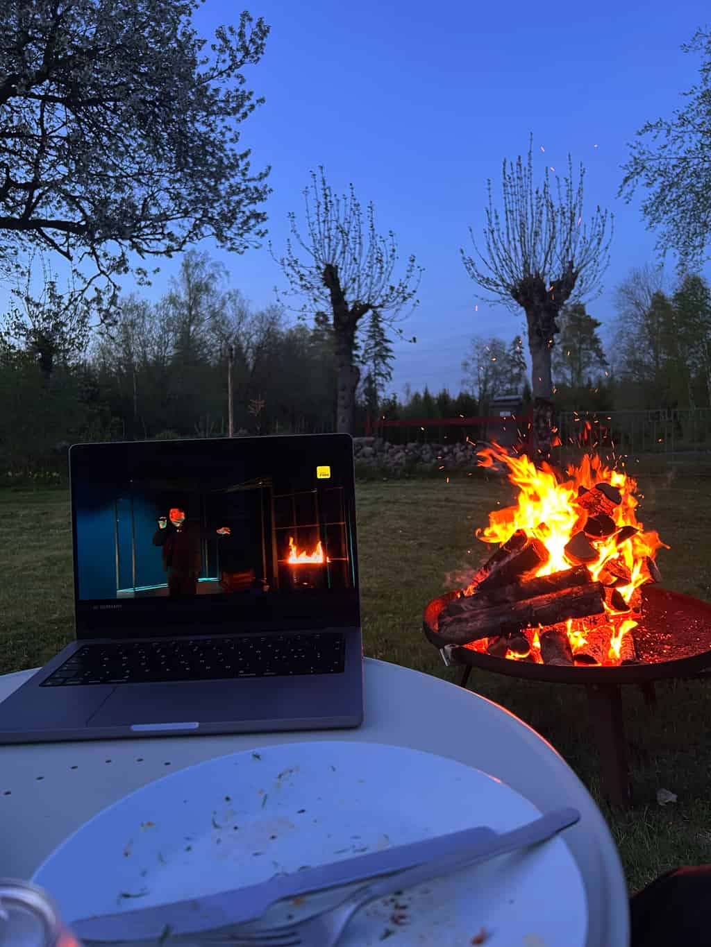 Ein Laptop, der im Freien auf einem Tisch steht, zeigt eine TV-Sendung, während ein Lagerfeuer im Hintergrund brennt.
