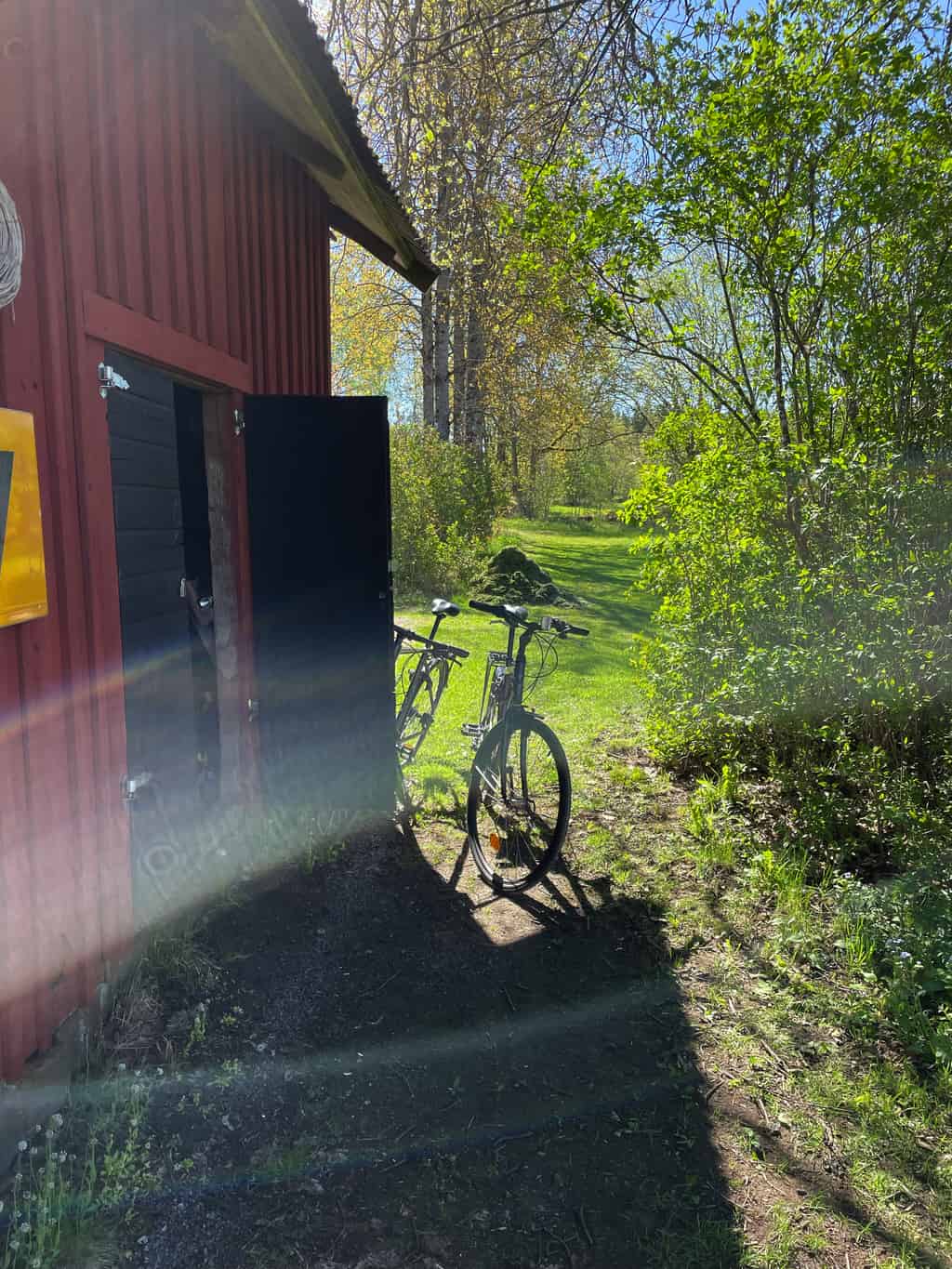 Zwei Fahrräder, die an der Seite eines roten Holzschuppens lehnen, mit grünen Bäumen und Rasen im Hintergrund.