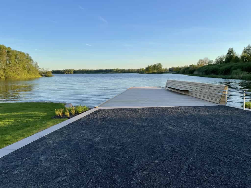 Ein Holzsteg, der zu einem ruhigen See führt, umgeben von grünen Bäumen und blauem Himmel.