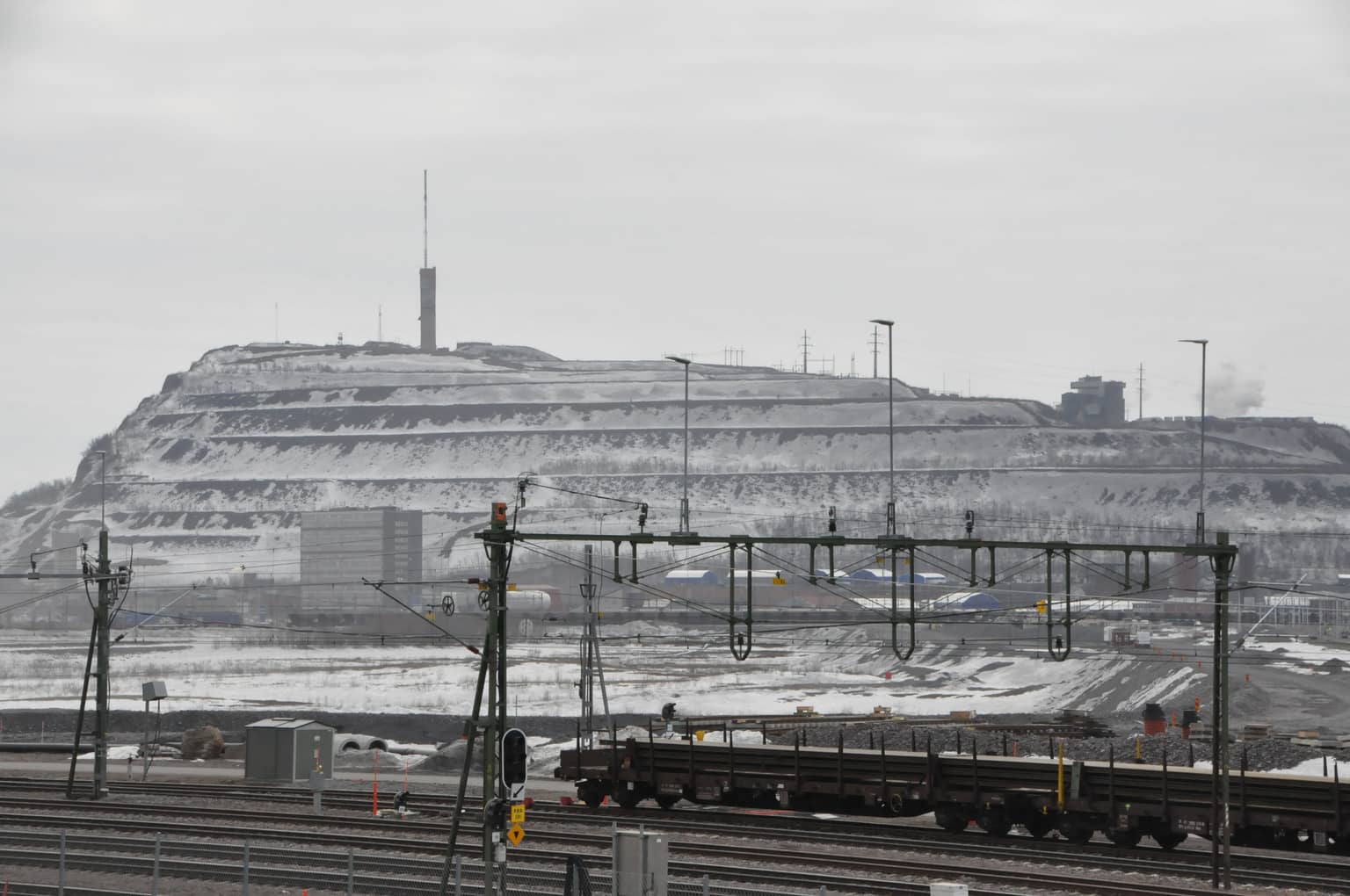 Weitblick auf eine verschneite Industrielandschaft mit einem großen Hügel, Bahngleisen und mehreren Industriegebäuden.