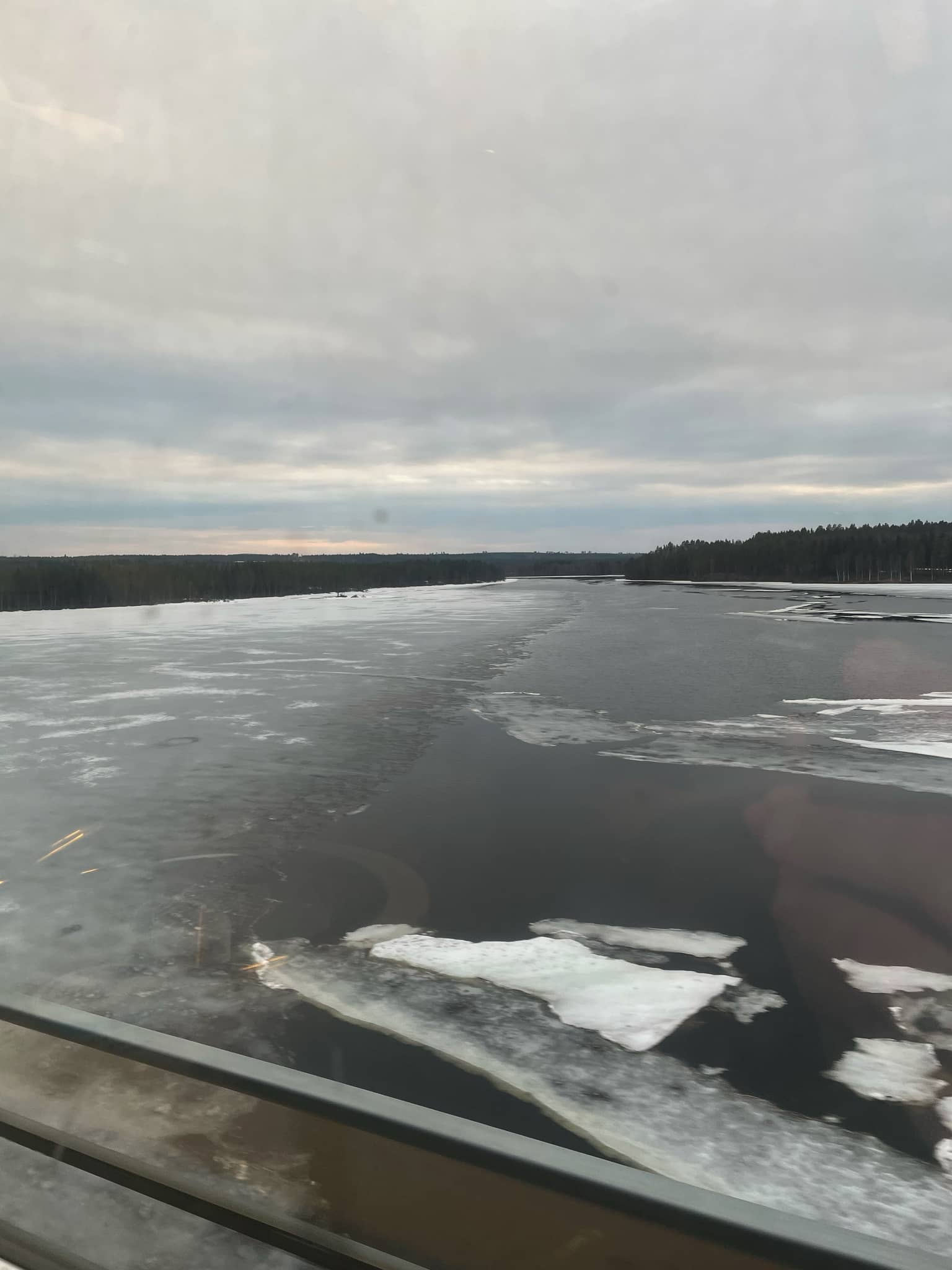 Blick aus einem Zugfenster auf einen zugefrorenen See, umgeben von bewaldeten Gebieten unter bewölktem Himmel.
