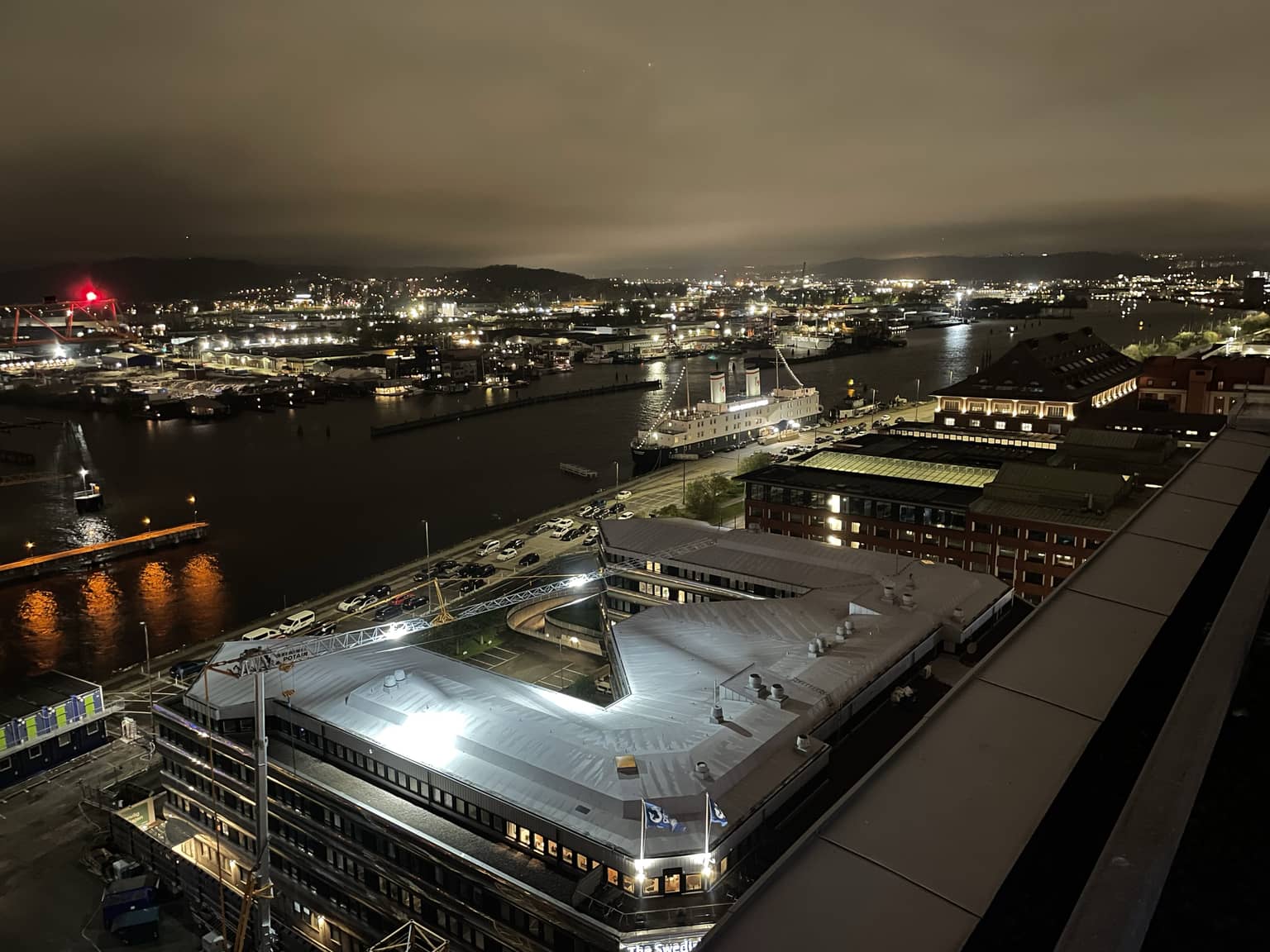 Nachtansicht eines beleuchteten Hafens mit einer leuchtenden Gebäudefassade im Vordergrund und der beleuchteten Stadt im Hintergrund, reflektiert auf dem Wasser.