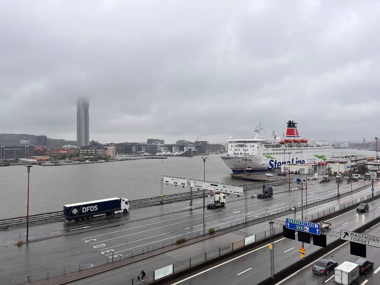 Blick auf eine Hafenszene an einem regnerischen Tag mit einem nebligen Hintergrund. Ein Schiff und mehrere Lastwagen sind sichtbar, und im Hintergrund ragt ein hohes Gebäude aus dem Nebel heraus.
