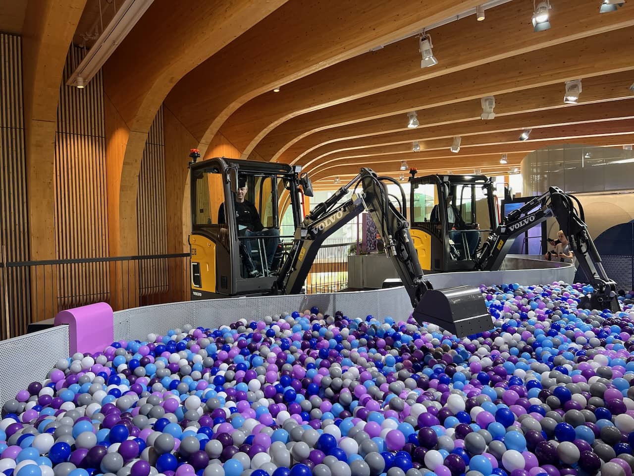 Zwei Bagger in einem Indoor-Spielbereich, gefüllt mit blauen und violetten Bällen, in einer Halle mit gewölbten Holzdecken.