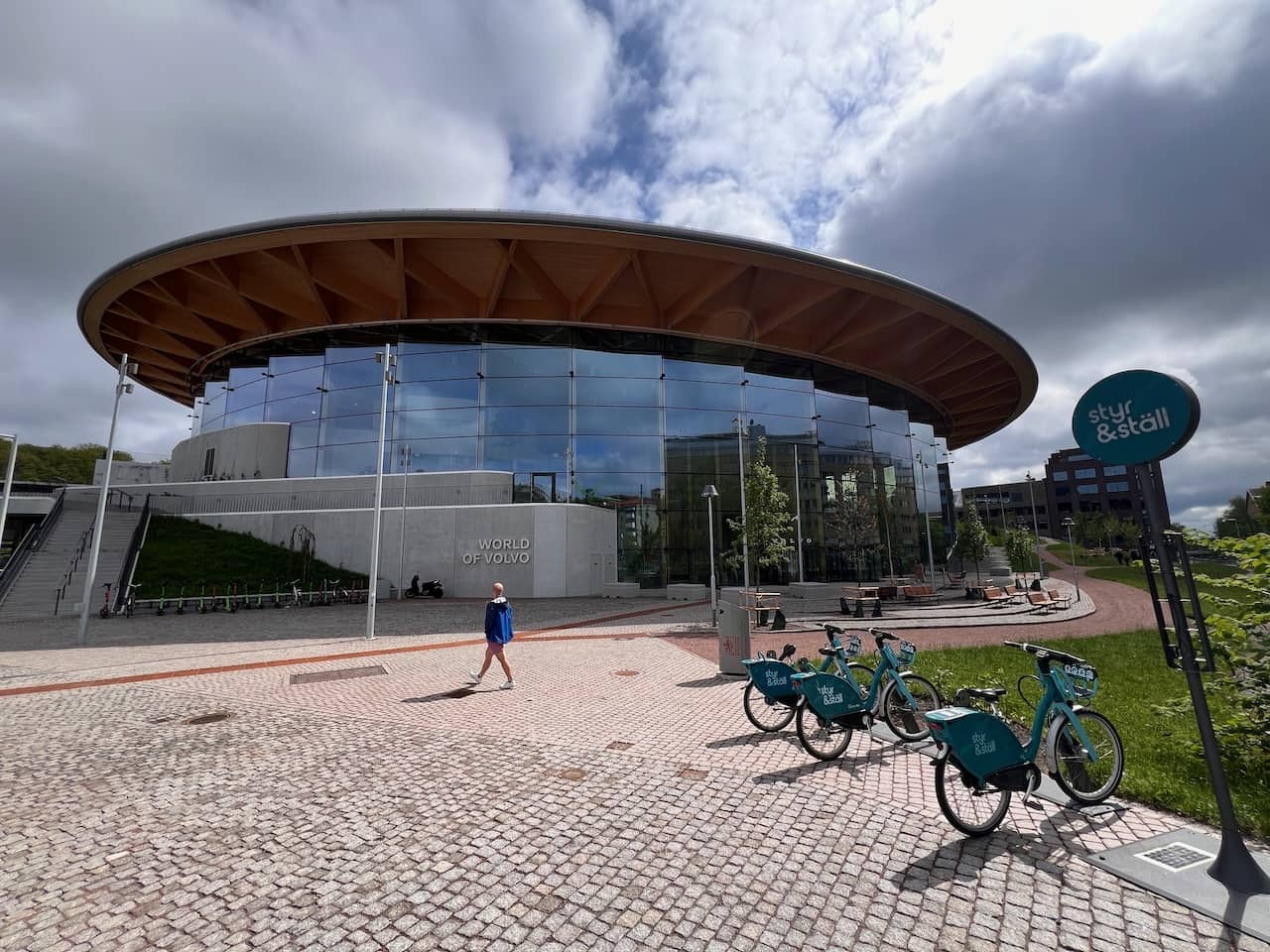 Das Gebäude 'World of Volvo' mit einer großen, kreisförmigen Holzstruktur und großen Glasfronten unter einem bewölkten Himmel. Im Vordergrund sind mehrere Mietscooter geparkt.