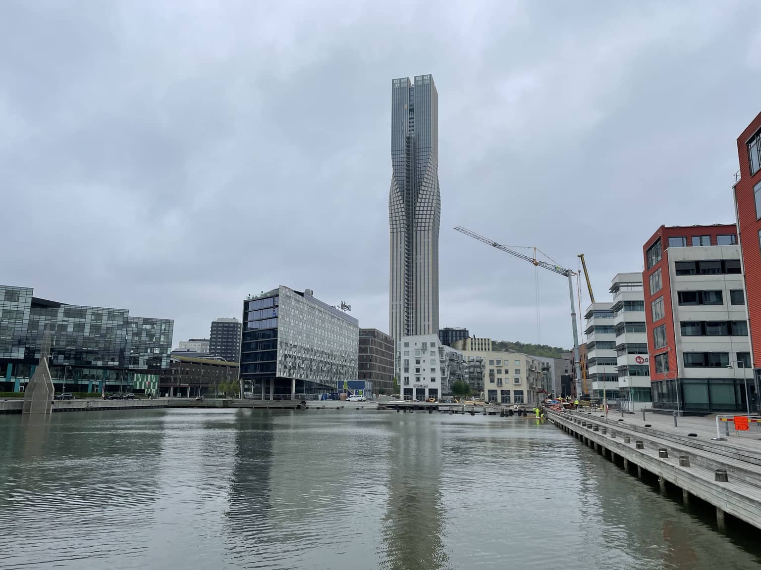 Blick auf das Ufer eines Flusses mit modernen Gebäuden und einem hohen, schlanken Turm. Das Bild zeigt eine städtische Umgebung mit einer reflektierenden Wasseroberfläche.