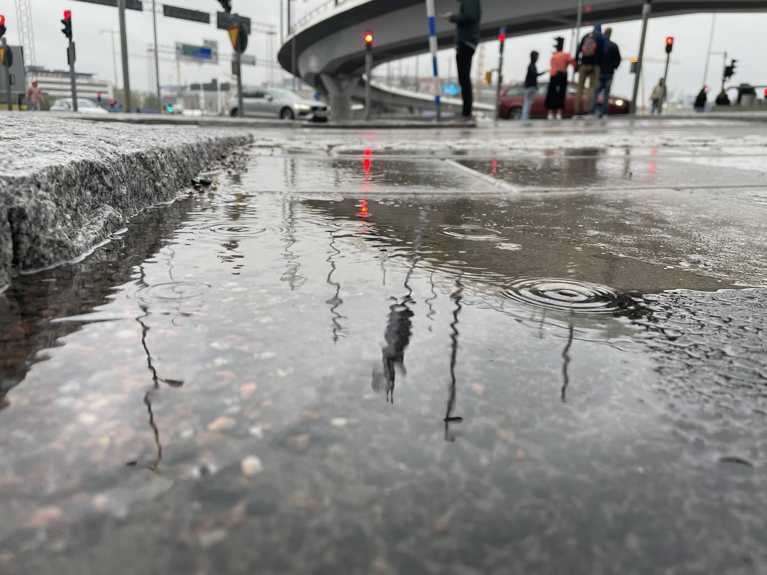 Eine städtische Szene an einem regnerischen Tag zeigt Pfützen auf dem Boden, die die Umgebung und Passanten spiegeln.