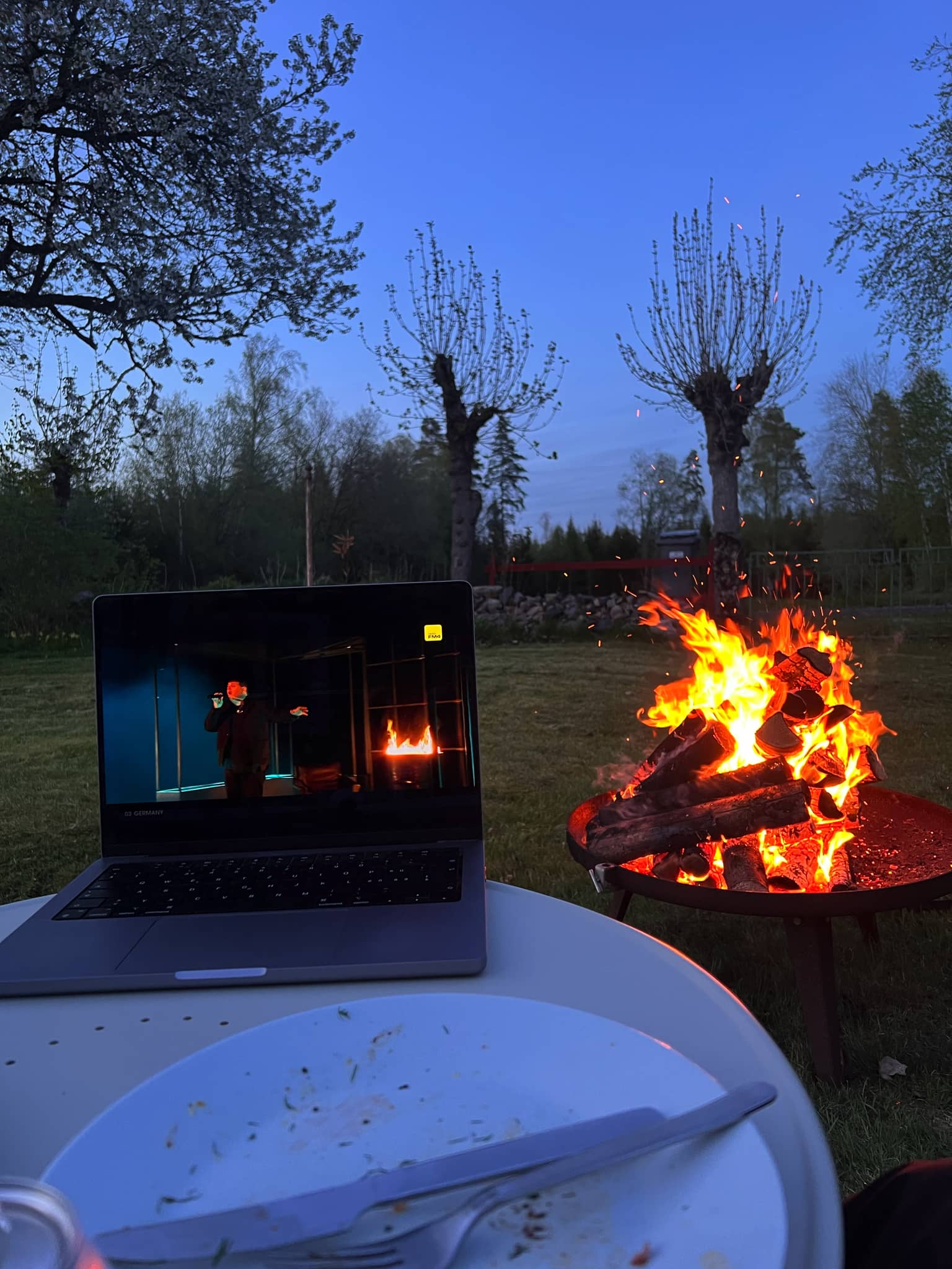 Ein Laptop, der im Freien auf einem Tisch steht, zeigt eine TV-Sendung, während ein Lagerfeuer im Hintergrund brennt.